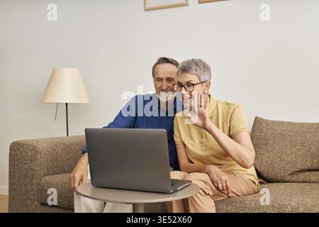 Cheerful senior couple using a laptop for video calling from the living room. Woman and man sitting Stock Photo