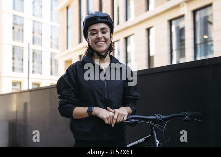 Young cheerful woman in cycling helmet standing in the city with a bike and looking at camera Stock Photo