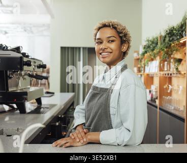 Young smiling barista in an apron leaning counter in a coffee shop. Portrait of a cheerful female bartender Stock Photo