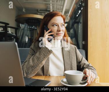 Beautiful businesswoman with ginger hair talking on smartphone and looking at window at a cafe Stock Photo