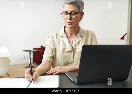 Mature woman writing on a note pad while using laptop computer at home Stock Photo