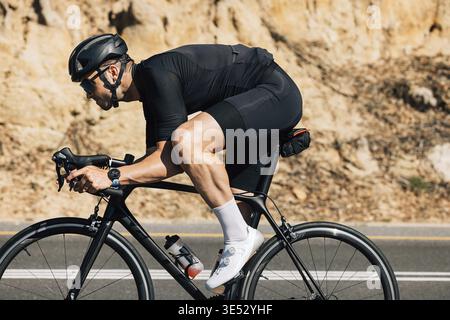 Professional cyclist in black sportswear riding road bike against the mountain. Muscular male doing intense training on bike outdoors Stock Photo