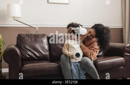 Young mother sitting on couch helping her son wear VR goggles Stock Photo