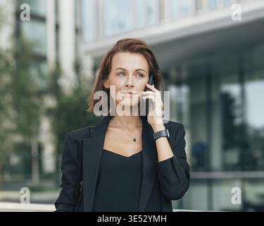 Middle-aged businesswoman in formal clothes making phone call. Portrait of a young adult female with ginger hair talking on a mobile phone while stand Stock Photo