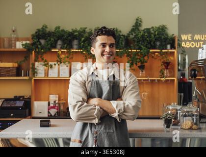 Confident barista in an apron. Young guy working as a barista in a coffee shop standing with crossed hands and looking away Stock Photo