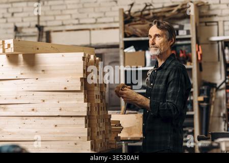 Senior man standing near a bunch wooden parts of furniture in a workshop and looking away Stock Photo