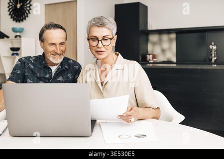 Senior retired couple at a table in dining room using a laptop computer. Two mature people looking at paperwork and discussing their finances Stock Photo