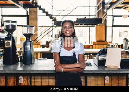 Portrait of a smiling coffee shop owner standing at counter wearing apron and arms crossed Stock Photo