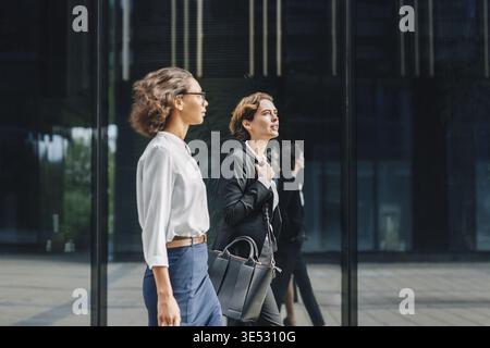 Two businesswomen walking to the office carrying her bags and talking Stock Photo