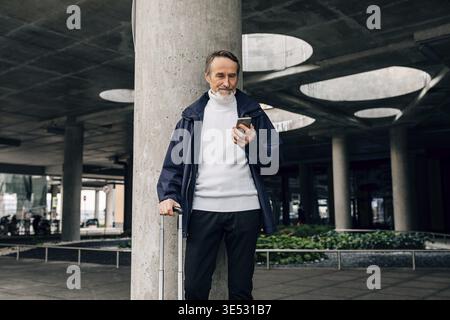 Senior man with rolling suitcase standing outdoors looking at mobile phone Stock Photo
