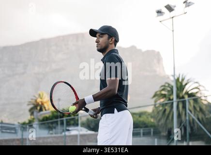 Fit man in sportswear and cap with a racket standing on tennis court. Tennis player looking away while standing on a hard court Stock Photo
