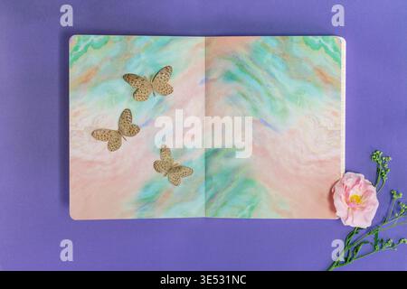 Pastel Marble Notebook with Decorative Butterflies and Pink Flower on Purple Background Stock Photo