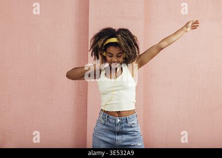 Cheerful woman with curly hair wearing yellow headphones dancing against a wall Stock Photo
