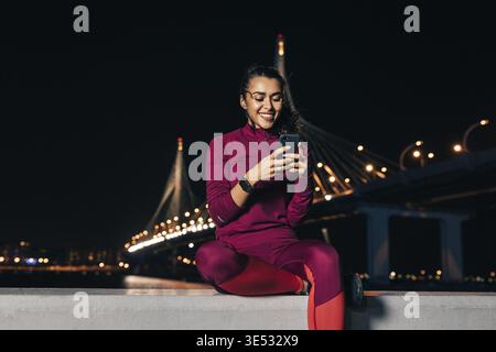 Smiling sportswoman sitting on an embankment using smartphone at night Stock Photo