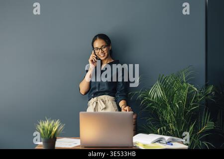 Smiling young woman standing at the table and looking on laptop screen while talking on cell phone Stock Photo