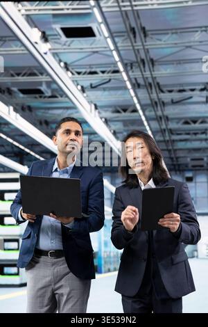 Multiethnic men work together in microchip factory, talking, comparing components production metrics. Teamworking management executives in PCB production plant analyze semiconductors operational data Stock Photo