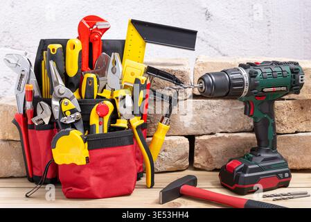Belt bag with various hand tools and cordless drill with drill bit next to an old brick on a wooden table. Selective focus. Stock Photo