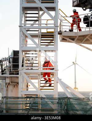 Offshore workers in orange PPE and hard hats climb the external staircase of a service operations vessel with a wind turbine visible behind, North Sea Stock Photo
