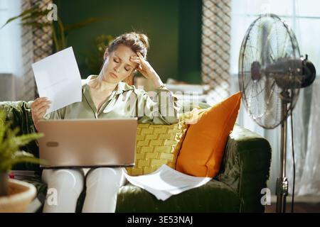 A mature woman on a green velvet couch looks exhausted, fanning herself with paper while working on a laptop. This scene captures the struggle of work Stock Photo