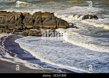 Powerful ocean waves gently breaking on the dark sandy beach and rugged rocky shoreline, creating white foam as they recede under the bright sunlight Stock Photo