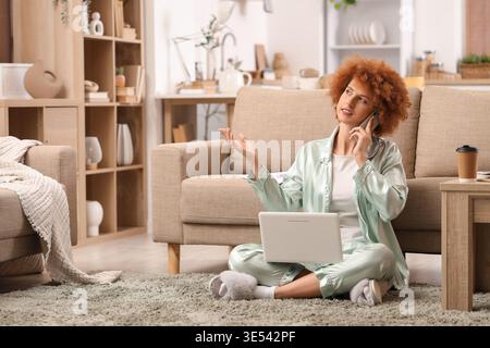 Redhead female freelancer with laptop talking by mobile phone at home Stock Photo
