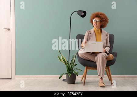 Redhead female freelancer working with laptop in armchair near green wall Stock Photo
