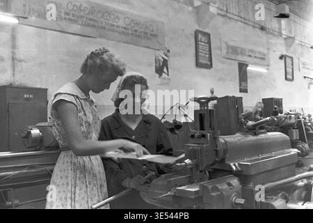 Sloviansk, 1950s-1960s, Armature-Insulator Plant (AIZ). A black and white historical photograph featuring a skilled female engineer in a light summer dress actively explaining technical blueprints to a dedicated female machinist. They stand closely together at a heavy metalworking machine, collaborating on specific manufacturing details under Soviet propaganda posters. This engaging archival image highlights women's professional roles in engineering and production, honoring the industrial heritage of the peaceful Donbas. Stock Photo