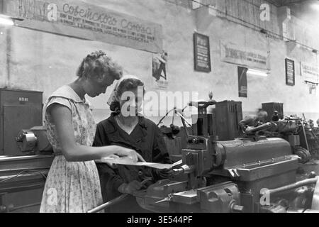 Sloviansk, 1950s-1960s, Armature-Insulator Plant (AIZ). A black and white historical photograph featuring a skilled female engineer in a light summer dress actively explaining technical blueprints to a dedicated female machinist. They stand closely together at a heavy metalworking machine, collaborating on specific manufacturing details under Soviet propaganda posters. This engaging archival image highlights women's professional roles in engineering and production, honoring the industrial heritage of the peaceful Donbas. Stock Photo