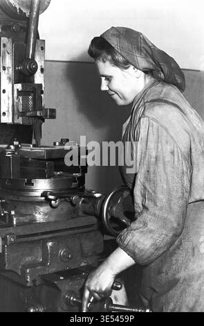 Sloviansk, 1950s-1960s, Armature-Insulator Plant (AIZ). A black and white historical photograph focusing on the profile of a focused female machinist operating a heavy industrial metalworking machine. Dressed in a protective headscarf, she carefully turns a lower control handwheel to adjust the mechanical equipment on the busy shop floor. This detailed archival image provides a realistic glimpse into Soviet factory environments, preserving the rich labor history and the manufacturing heritage of the peaceful Donbas. Stock Photo