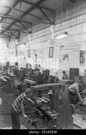 Sloviansk, 1950s-1960s, Armature-Insulator Plant (AIZ). A black and white historical photograph providing a comprehensive view of a bustling mechanical workshop. Numerous skilled male workers are actively operating heavy metalworking milling machines and lathes. The bright industrial interior is decorated with authentic Soviet propaganda posters and motivational slogans on the walls. This incredibly detailed archival image preserves the intense labor history and the heritage of the peaceful Donbas. Stock Photo