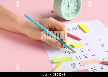 Female hand with alarm clock, pencils and sticky notes marking date in calendar on pink background. Planning concept Stock Photo