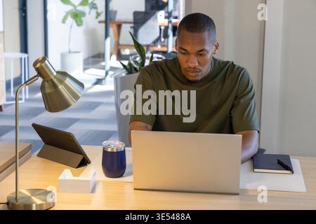 African American man wearing olive tee working on silver laptop at office desk, tablet nearby Stock Photo