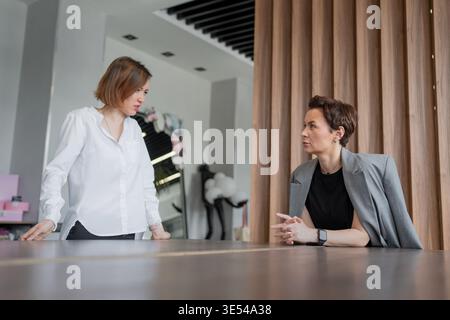 Women talking and collaborating while standing and sitting at table in modern bright business office. Collaboration, talking, teamwork, office, women. Stock Photo