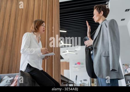 Businesswomen standing and talking in front of wood wall inside modern company office. Business, standing, talking, teamwork, office, women. Stock Photo