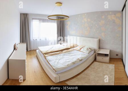 A contemporary bedroom interior with wood paneling, beige bed, armchair, pendant lights, soft curtains, and herringbone parquet flooring. Stock Photo