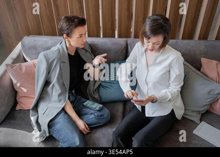 Female coworkers in casual business clothing talking candidly together on sofa in modern open office. Coworkers, business, candid, sofa, teamwork Stock Photo