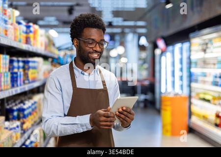 African american male employee wearing an apron and eyeglasses standing inside a grocery store aisle, using a digital tablet to manage inventory and check stock, smiling as he works Stock Photo