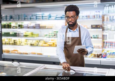 African american man wearing an apron and glasses standing in a supermarket aisle, holding a tablet computer while checking inventory in a frozen food freezer, working efficiently to manage stock Stock Photo