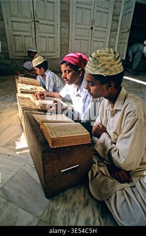 Students are focused on reading and studying from Quran books in a madrasa. The room has simple furniture and is filled with natural light. Stock Photo