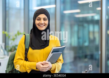 Muslim student woman wearing a hijab and backpack, confidently smiling while holding notebooks, representing education, success, diversity, and modern learning in a blurred university setting Stock Photo