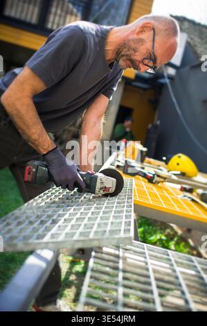 A man is using a power tool to cut a metal grid in an outdoor workshop. Various tools are scattered around him on a workbench. The sun is shining on t Stock Photo