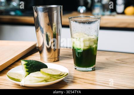 Mixing glass containing green liquid and cucumber slices sits on wooden counter next to metal shaker and lime. cucumber lime mocktail, refreshing Stock Photo