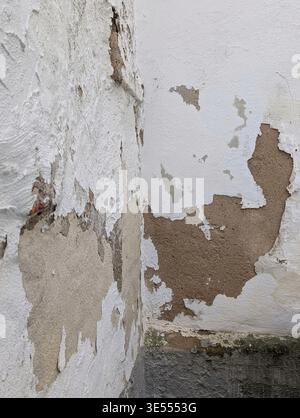 Peeling Plaster Corner Wall Revealing Brown Underlayer, Flaking Paint And Damp Stains Along Rough Concrete Base, Muted Beige And White Tones, Textured Stock Photo