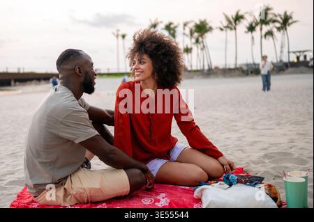 Casual young couple enjoying a spontaneous evening picnic on the sand focused on their relaxed style Stock Photo