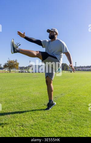 Adult African American man performing kick, balancing on one leg on field, wearing cap and sneakers Stock Photo