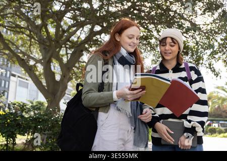 Female students standing under large tree on campus walkway, studying open notebook and gray binder Stock Photo