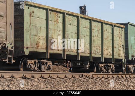 Rusty green freight railway cars on tracks, closeup view of weathered metal panels, wheelsets and gravel ballast, warm midday light, industrial siding Stock Photo