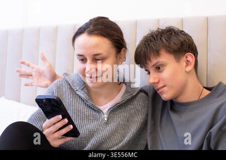 Happy teen siblings relaxing on sofa, looking at smartphone and smiling. Cozy home scene with digital communication, bonding and shared leisure time. Stock Photo