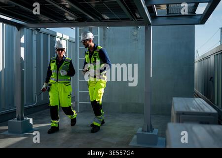 Male and female engineers inspecting photovoltaic systems beneath solar panel structures on a rooftop, representing professional renewable energy management || Model released Stock Photo