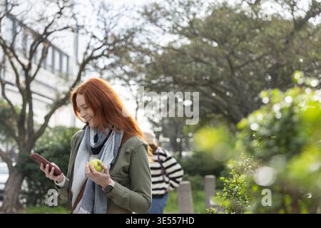 Red-haired woman standing in park wearing coat, scarf, smartwatch, holding apple, checking phone. Urban, greenery, smartphone, casual, candid, daylight, trees || Model released Stock Photo